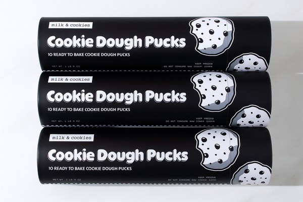 Three black cylindrical packages of milk & cookies Cookie Dough Pucks stacked vertically, each containing 10 ready-to-bake pucks with chocolate chip cookie illustrations.