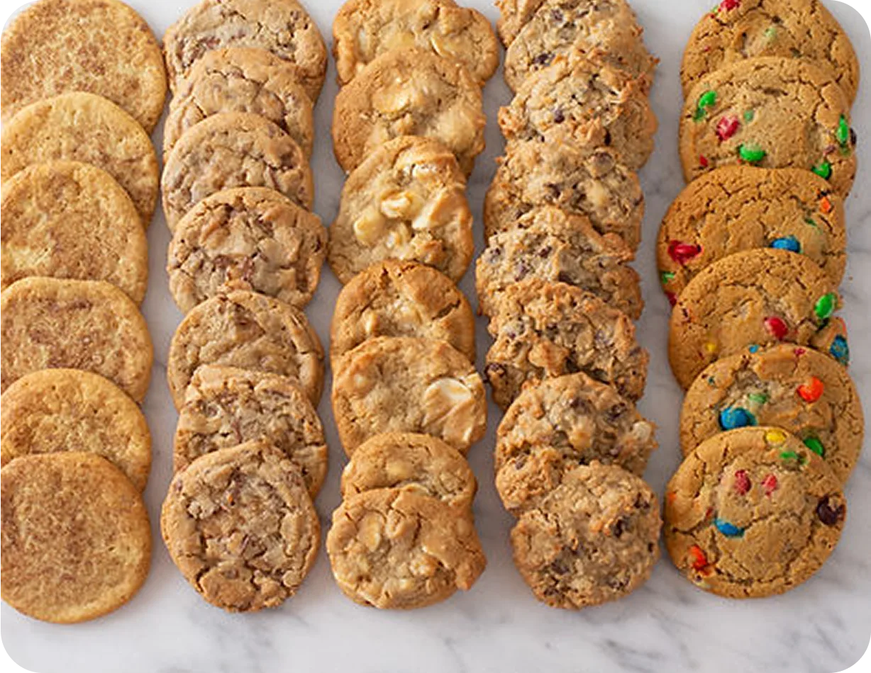 Five rows of assorted cookies including snickerdoodle, chocolate chip, white chocolate macadamia nut, trail mix, and colorful M&M varieties arranged on white marble surface.