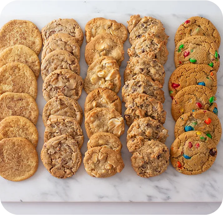 Five rows of assorted cookies including plain sugar, chocolate chip, white chocolate macadamia, oatmeal raisin, and colorful M&M cookies arranged on white parchment paper.