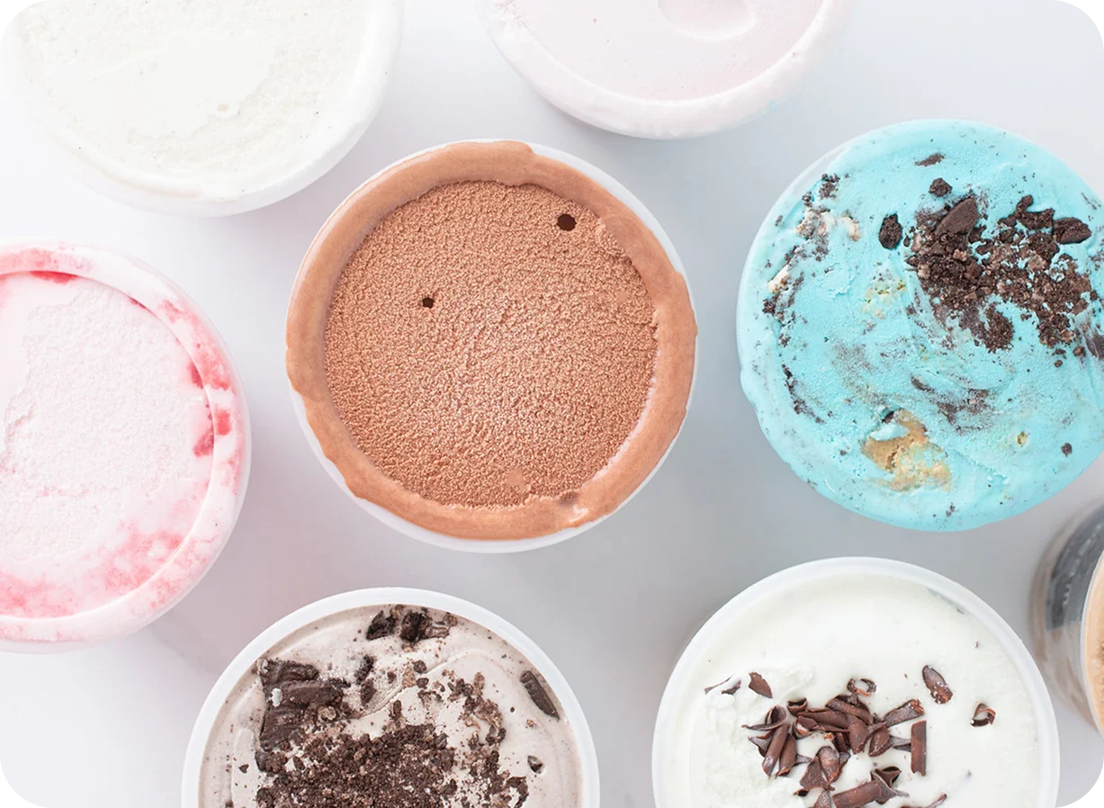 Various ice cream flavors in containers viewed from above, including chocolate, strawberry, mint chip, and vanilla with toppings