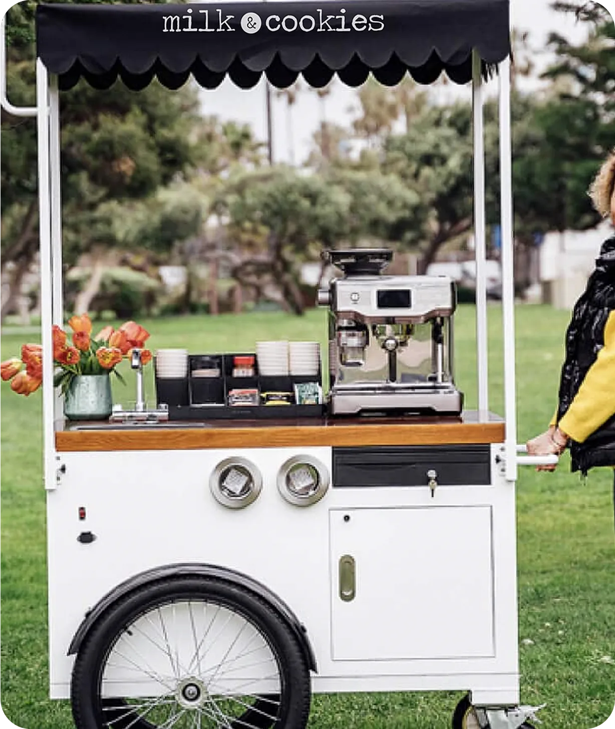 White mobile coffee cart with black canopy reading "milk & cookies" on grass, featuring espresso machine, flowers, and storage compartments
