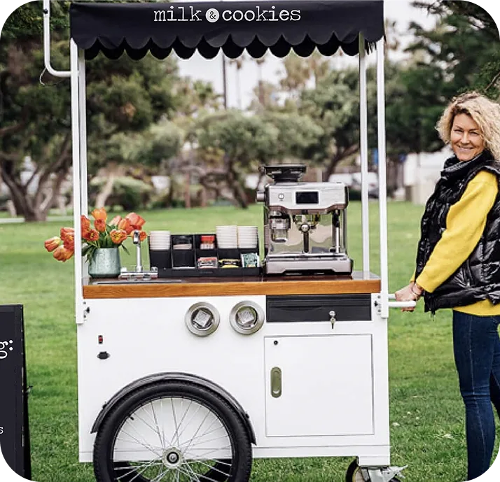 Woman in yellow shirt and black vest stands beside white mobile coffee cart with "milk & cookies" awning in grassy park setting