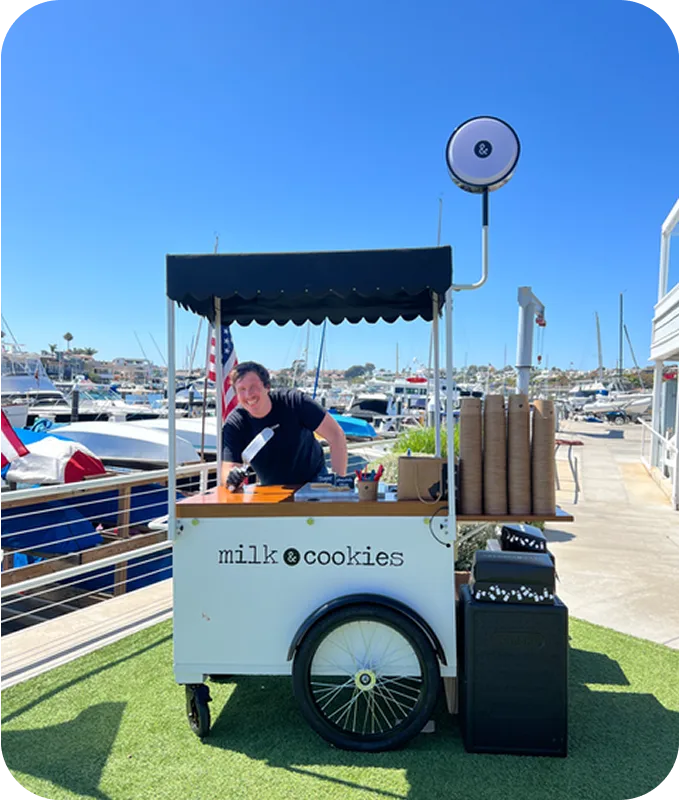 A vendor operates a white "milk cookies" cart with black canopy at a sunny marina with boats and blue sky in background.