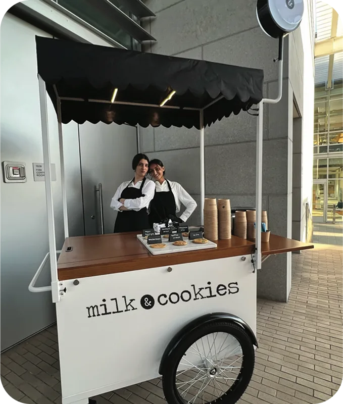 Two people in aprons standing behind a white mobile cart with black awning labeled "milk & cookies" on brick pavement
