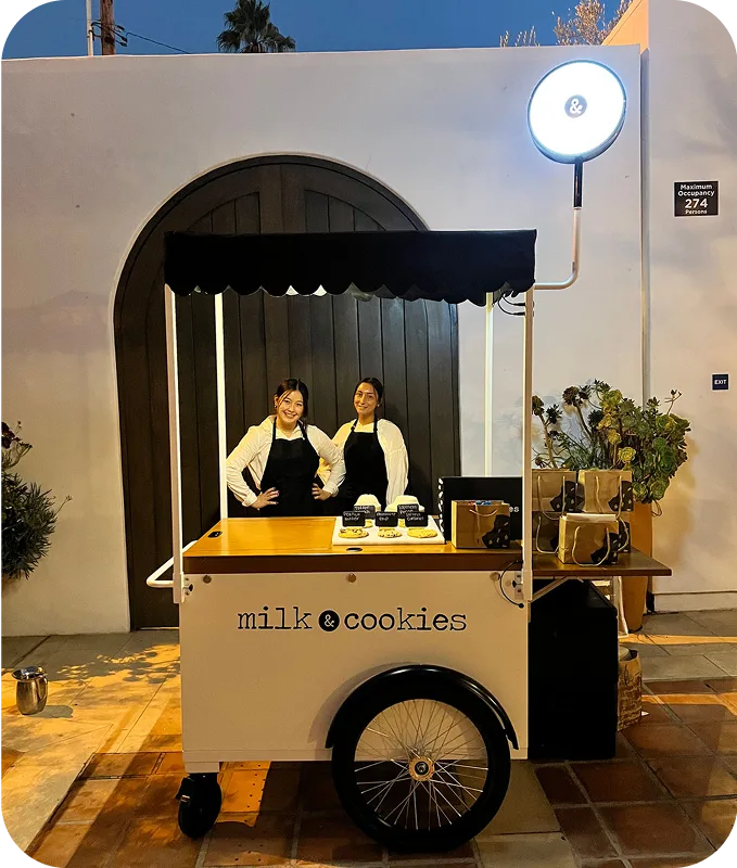 Two women in black aprons stand behind a white mobile milk & cookies cart with black wheels outside a modern white building with arched entrance.