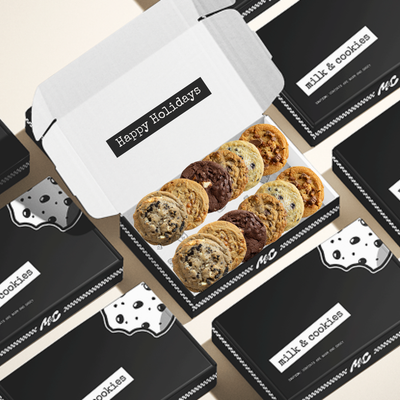 White gift box with "Happy Holidays" label containing assorted cookies, surrounded by black "milk & cookies" branded boxes