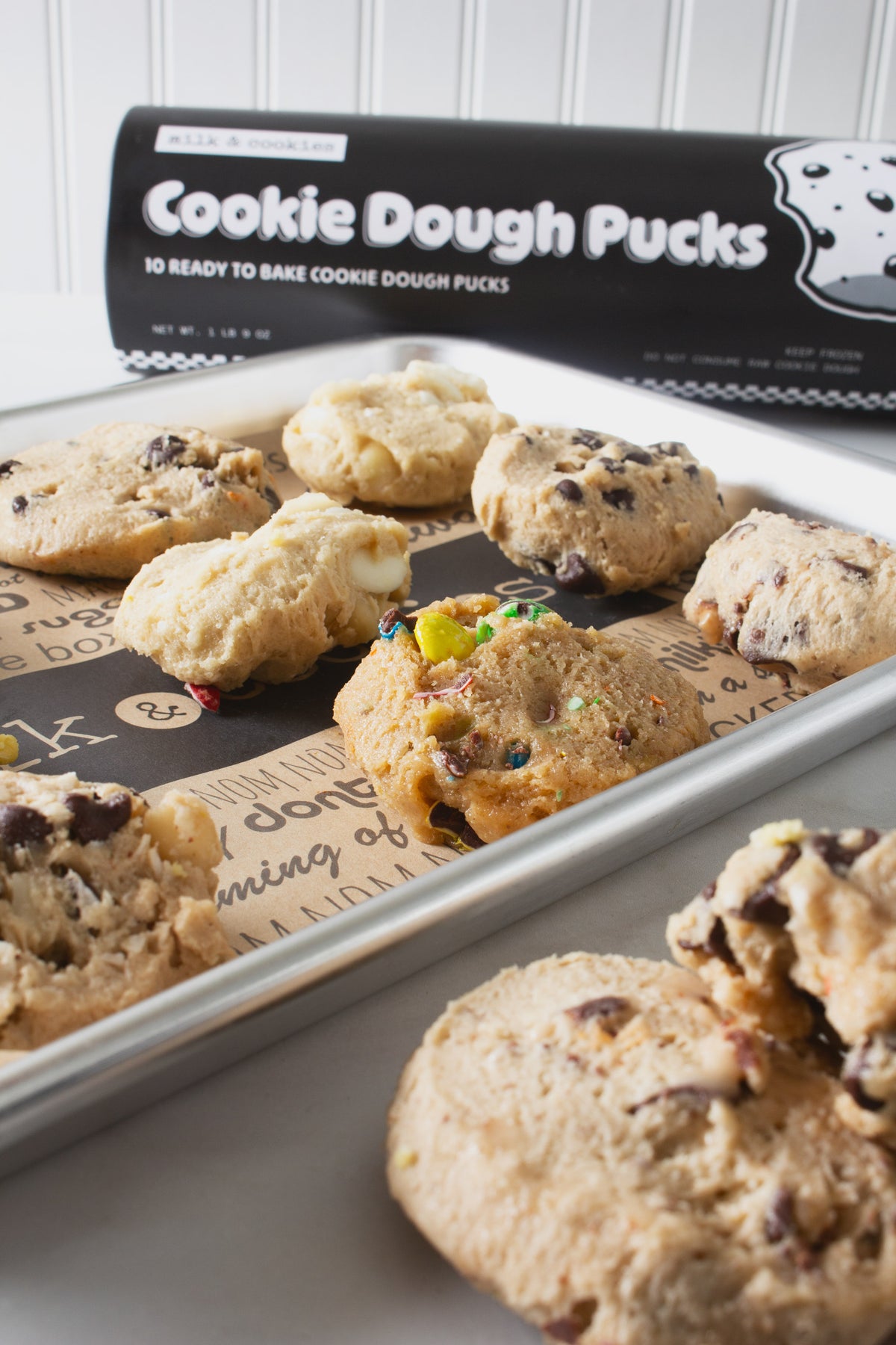 Cookie dough pucks with various mix-ins like chocolate chips and colorful candies arranged on a dark baking sheet, with black packaging visible in background.