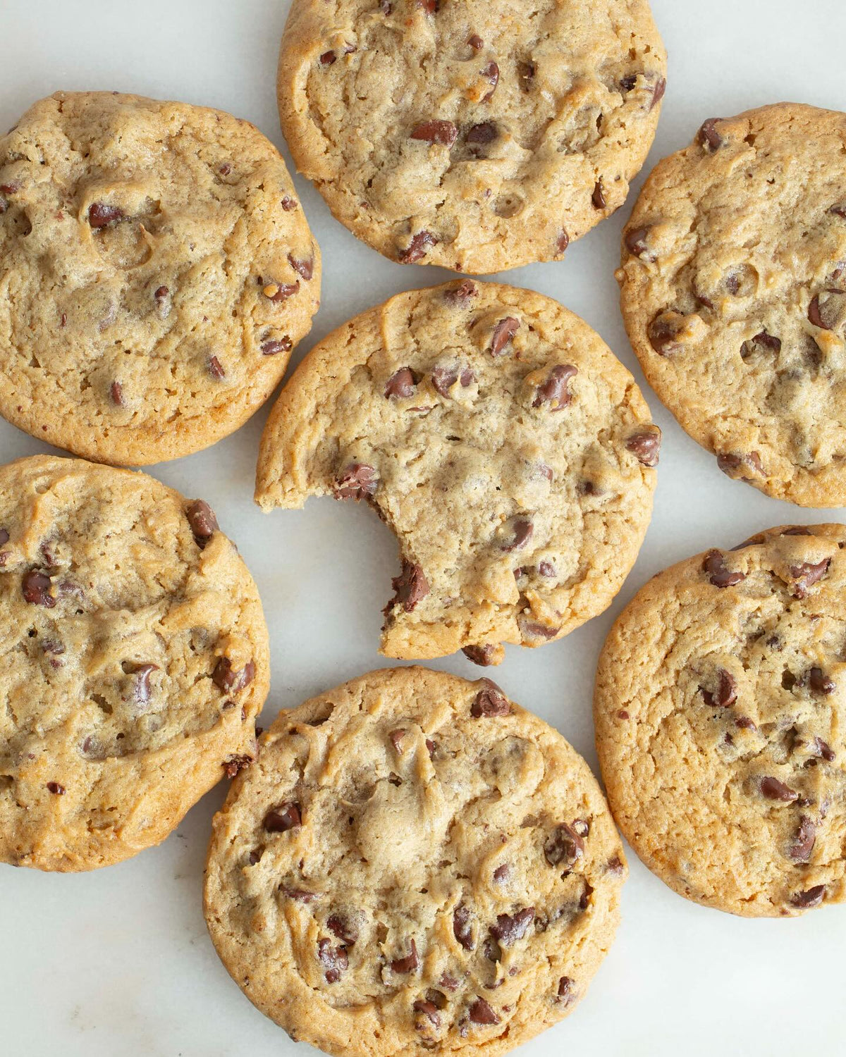 Golden brown chocolate chip cookies arranged on white surface, showing soft chewy texture with visible chocolate chunks and bite taken from one cookie
