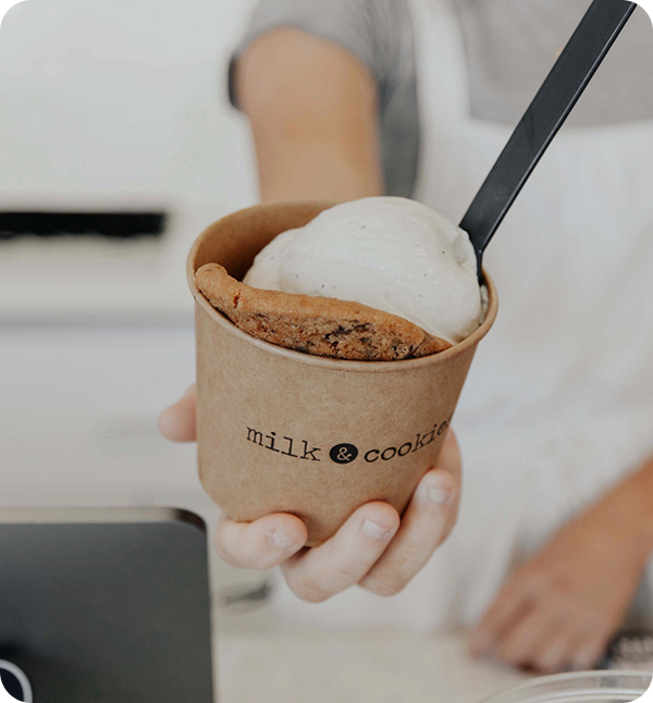 Person in white shirt holding brown paper cup with vanilla ice cream scoop and chocolate chip cookie, black spoon inserted